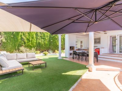 Terrasse extérieure couverte avec coin repas et barbecue intégré, jouxtant une pelouse artificielle dotée d'un salon de jardin et d'un grand parasol.