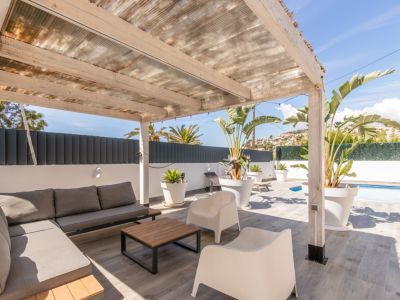 Terrasse extérieure couverte avec pergola en bois et toit translucide, aménagée d'un canapé modulaire gris, d'une table basse et de plantes tropicales. Sol carrelé et vue partielle sur la piscine.