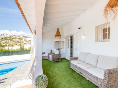 Vaste terrasse couverte meublée de canapé et fauteuils en osier sur gazon synthétique, avec vue sur la piscine privée et les collines résidentielles.