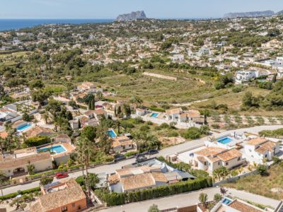 Vue aérienne d'un quartier résidentiel à Moraira, avec des villas aux toits en terre cuite et des piscines privées. La mer Méditerranée et un cap rocheux lointain encadrent ce paysage côtier verdoyant.