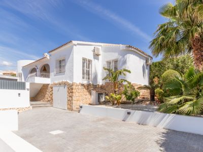 Vue de face de la villa avec façade blanche, soubassement en pierre naturelle et toit en tuiles. Une grande allée pavée et un jardin luxuriant avec palmiers sont visibles.