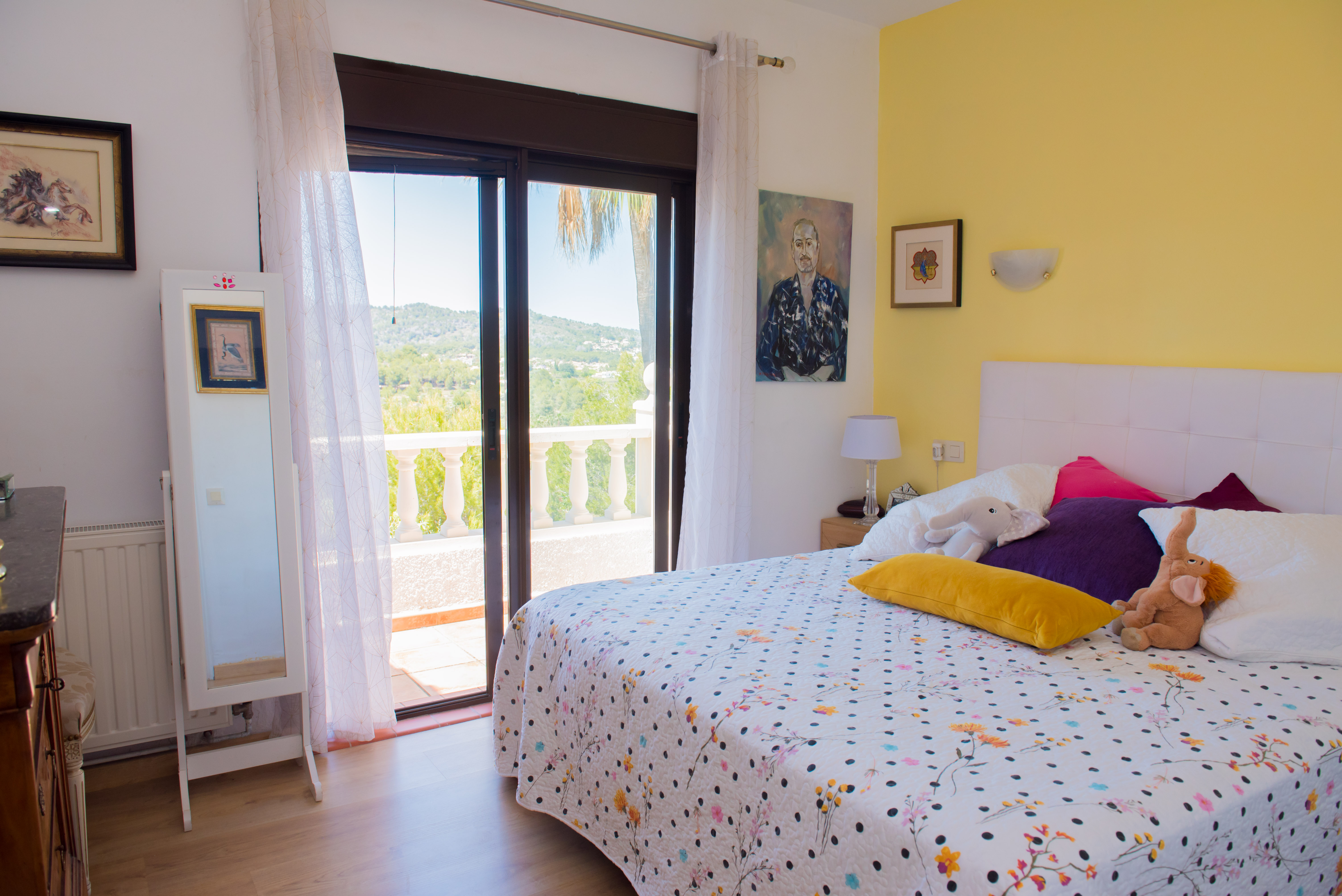 Chambre lumineuse avec accès au balcon. Mur jaune et literie à motifs floraux.