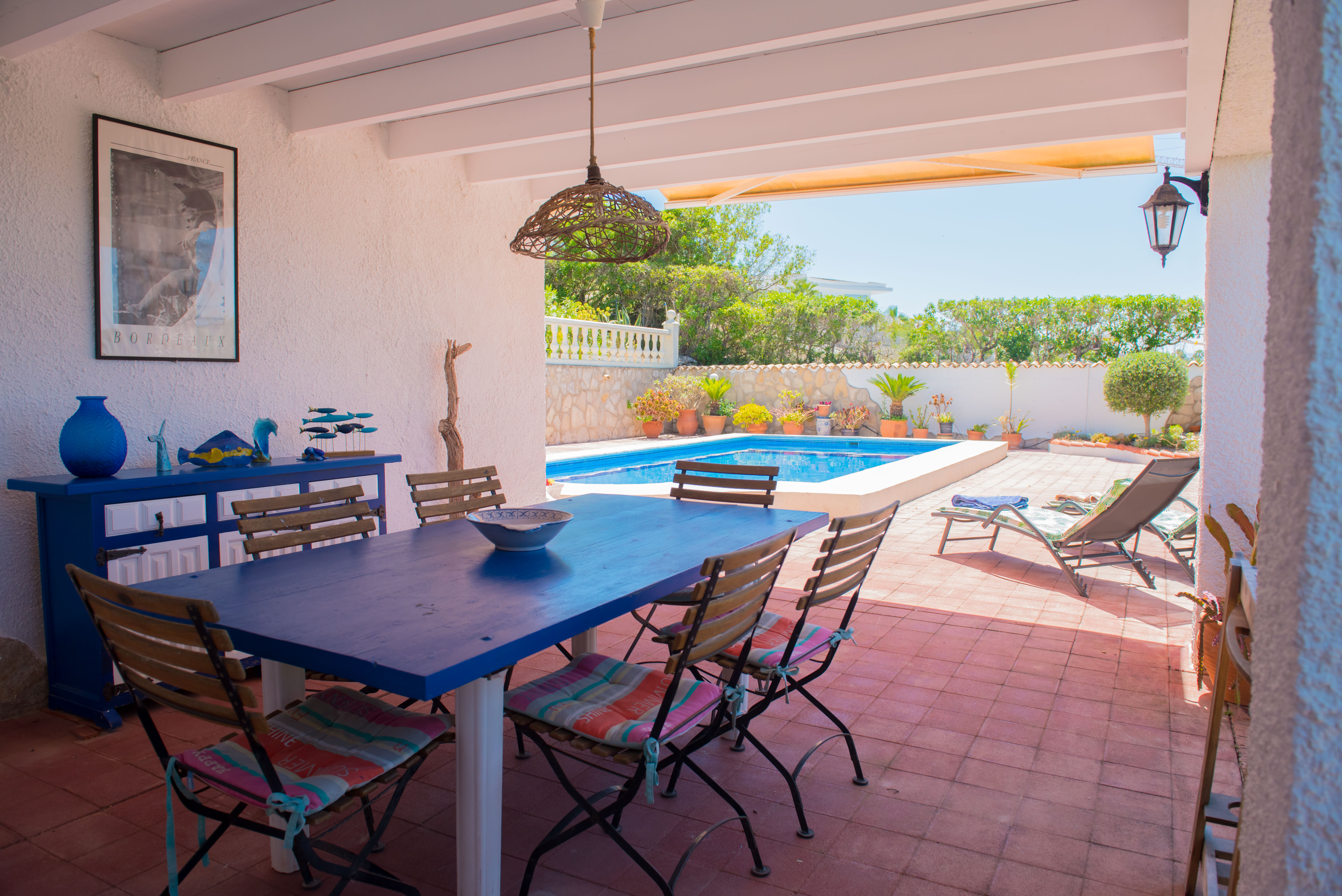 Salle à manger couverte avec table bleue et vue sur la piscine.