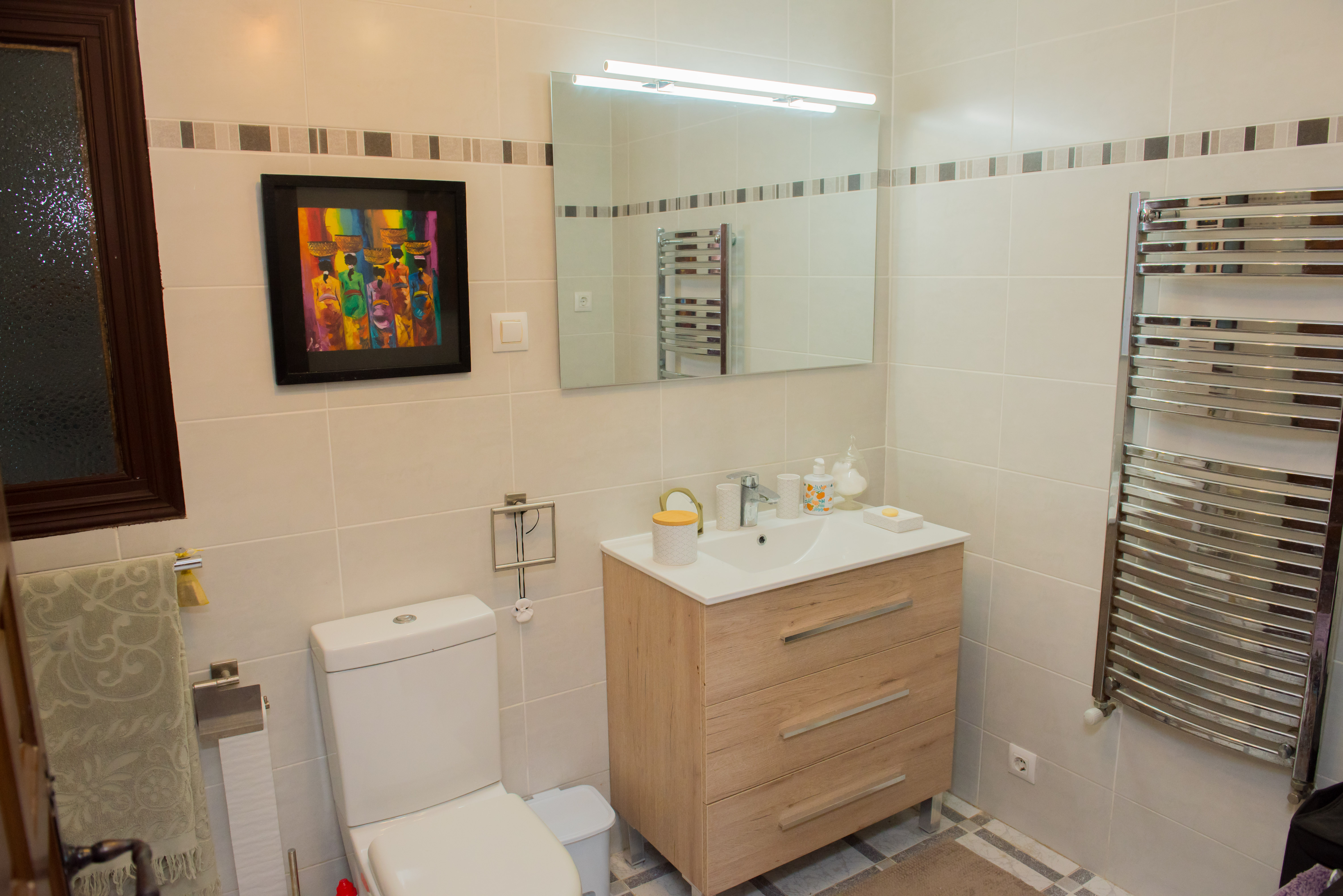 Salle de bain avec carrelage crème, meuble-lavabo en bois clair et sèche-serviettes chromé.