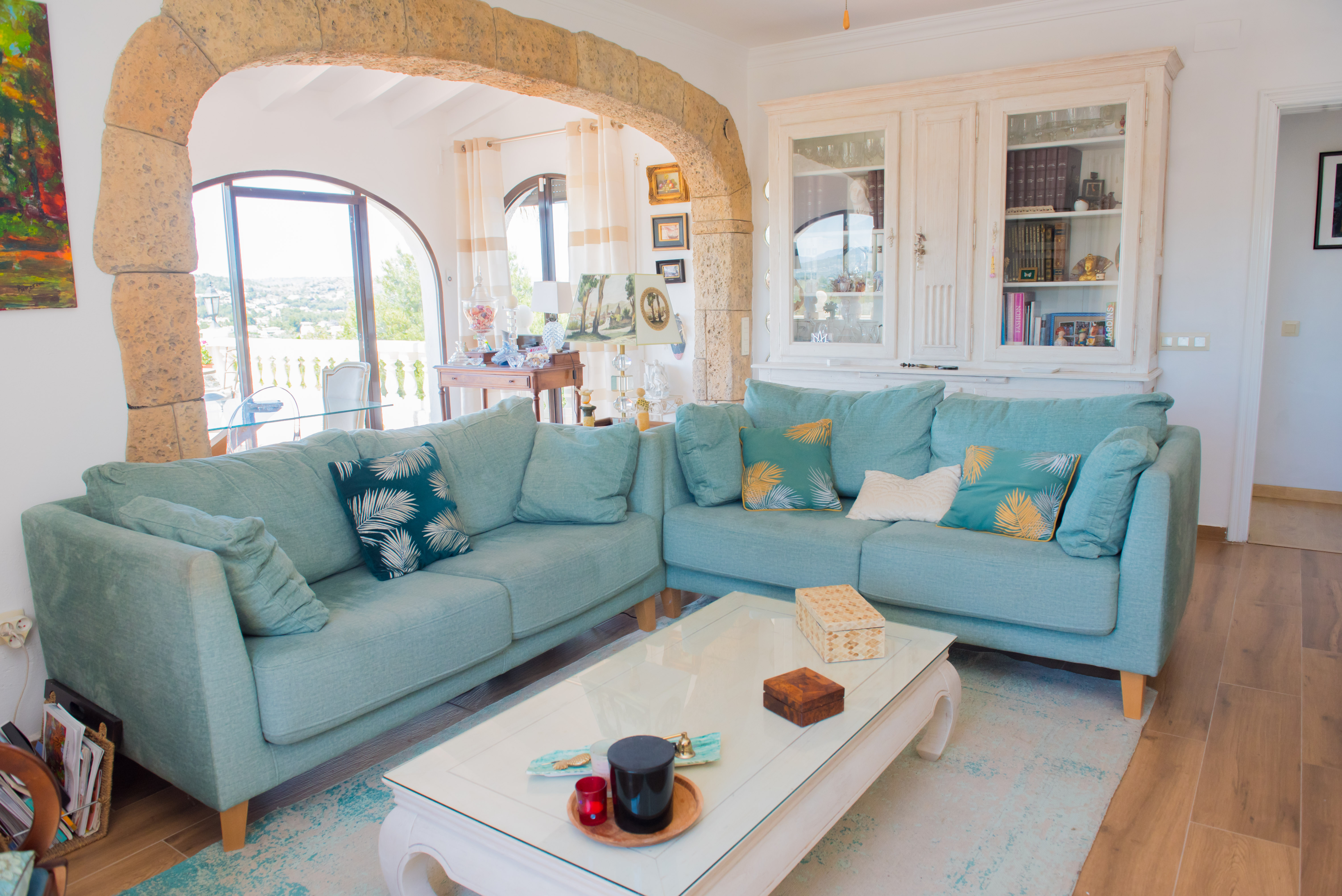 Salon lumineux avec canapé d'angle turquoise, table basse blanche et arche en pierre rustique encadrant la vue extérieure.
