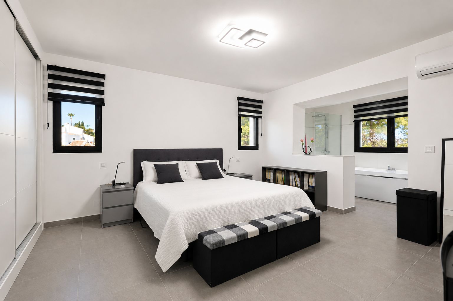 Chambre lumineuse avec carrelage gris, lit double avec tête de lit rembourrée et salle de bain attenante via une ouverture.