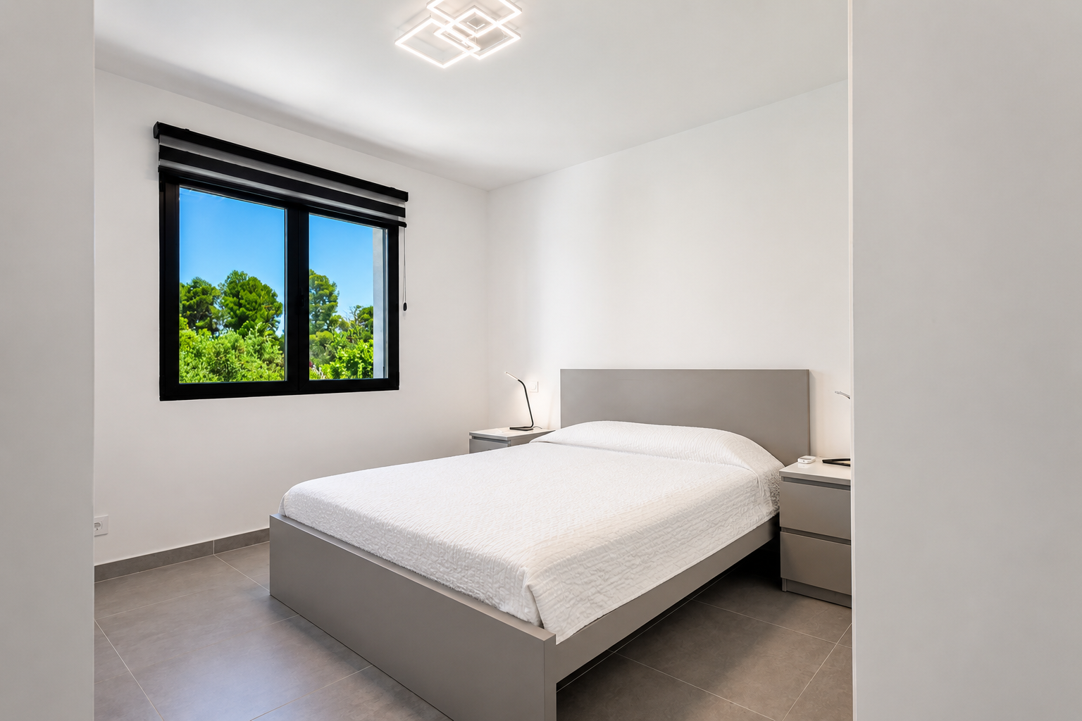 Chambre lumineuse avec lit double gris, éclairage moderne et vue sur le jardin à travers la fenêtre à cadre noir.