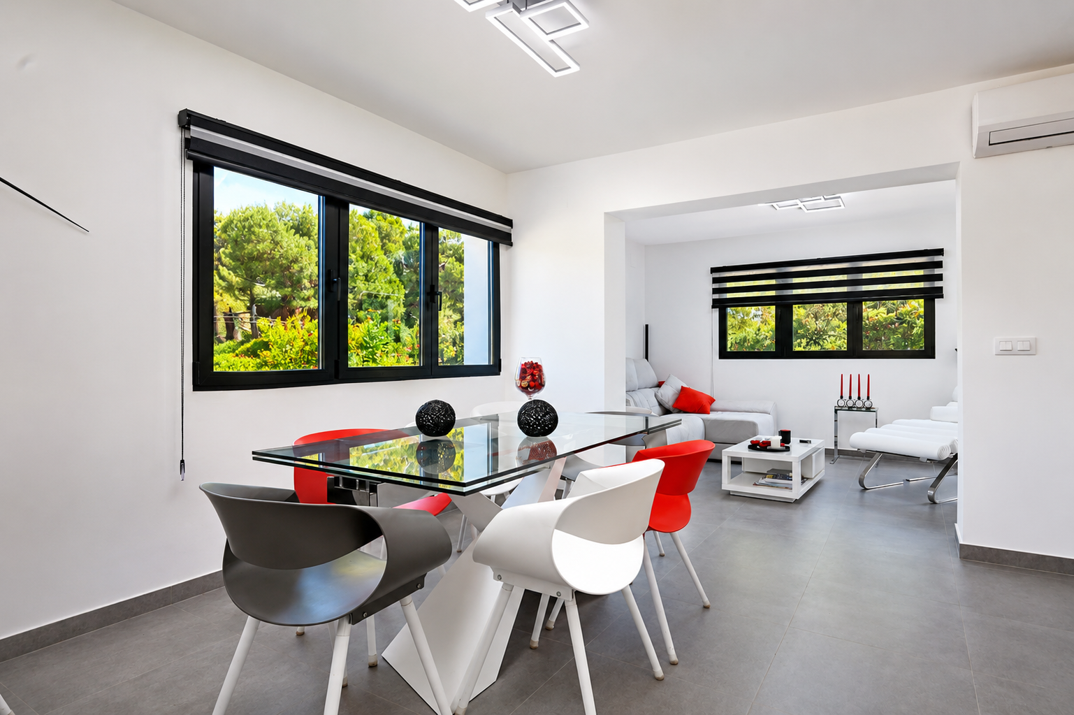 Salle à manger lumineuse avec table en verre, chaises design et vue sur le jardin à travers des fenêtres avec stores.