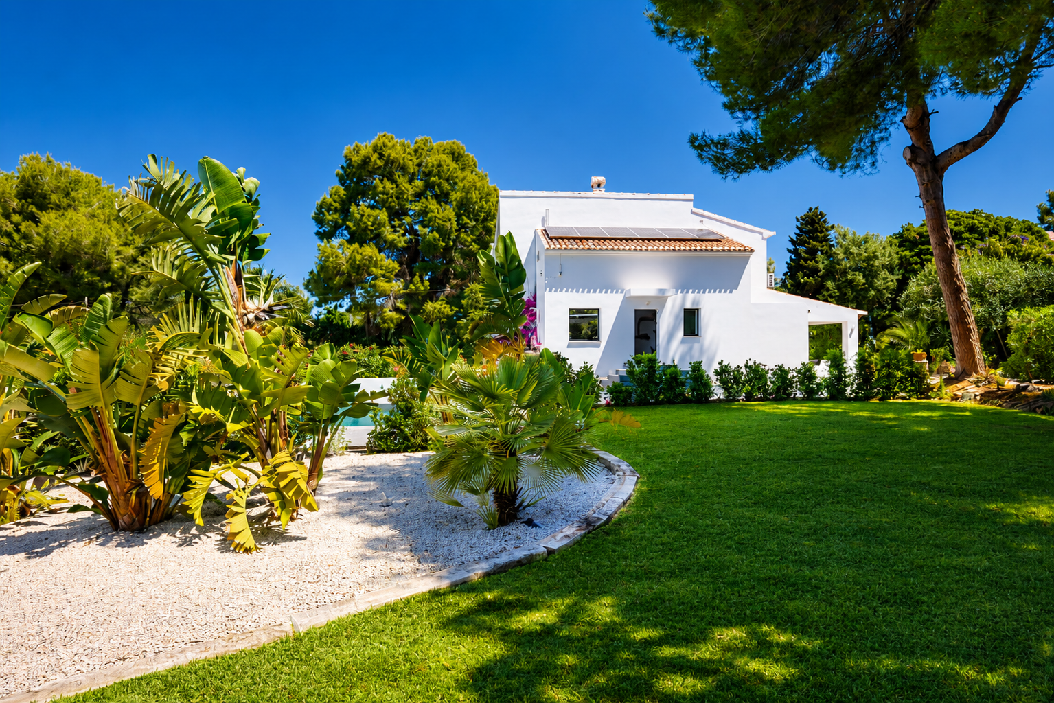 Vue extérieure de la villa avec façade blanche, jardin avec pelouse et végétation tropicale.