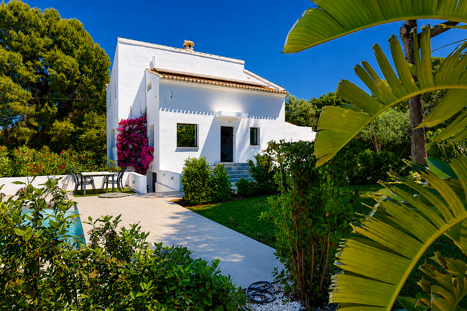 Vue extérieure de la villa blanche avec jardin, piscine et bougainvilliers sur la façade.