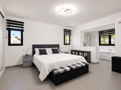 Chambre lumineuse avec carrelage gris, lit double avec tête de lit rembourrée et salle de bain attenante via une ouverture.