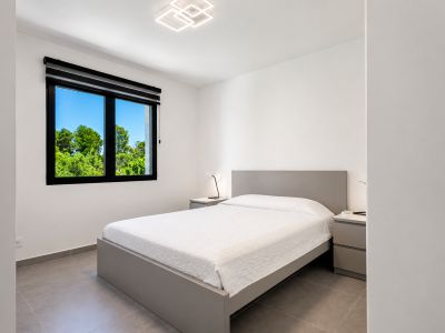 Chambre lumineuse avec lit double gris, éclairage moderne et vue sur le jardin à travers la fenêtre à cadre noir.