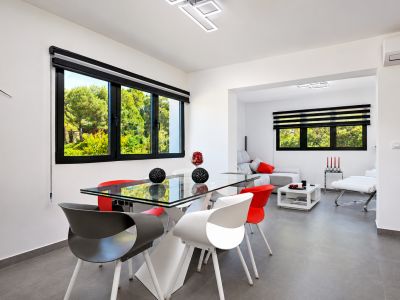 Salle à manger lumineuse avec table en verre, chaises design et vue sur le jardin à travers des fenêtres avec stores.