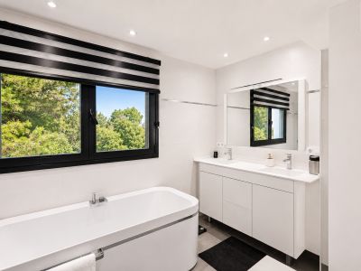 Salle de bain lumineuse avec fenêtre à cadre noir offrant une vue extérieure, baignoire blanche et meuble-lavabo double avec miroir.