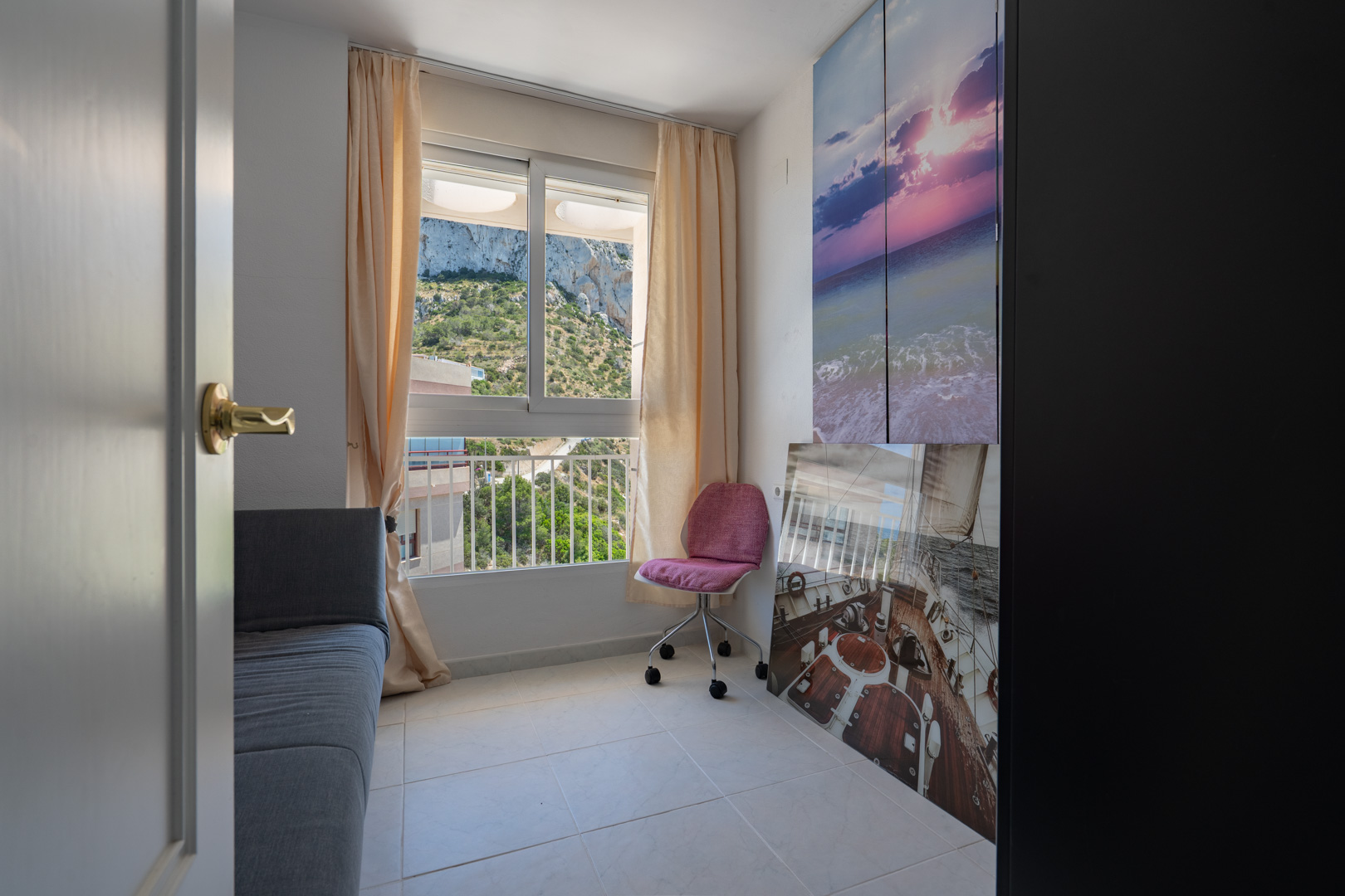 Cette pièce polyvalente dispose d'une fenêtre offrant des vues directes sur la colline verdoyante, complétée par un canapé-lit gris et une chaise de bureau rose. Des œuvres d'art murales ajoutent une touche côtière.