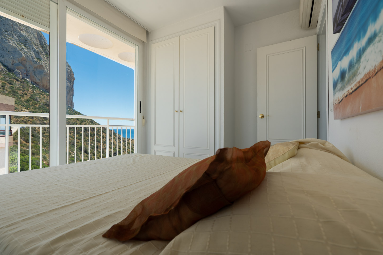 Chambre avec placards intégrés blancs et porte coulissante vitrée ouvrant sur un balcon privé offrant des vues directes sur le Peñón de Ifach et la mer.