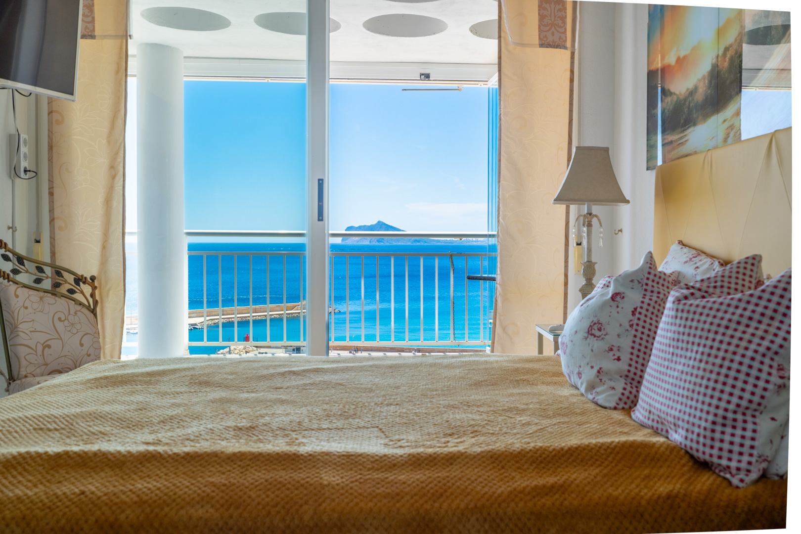 Chambre confortable avec lit et accès direct à un balcon, offrant des vues panoramiques sur la mer bleue, le port de plaisance et une île lointaine.