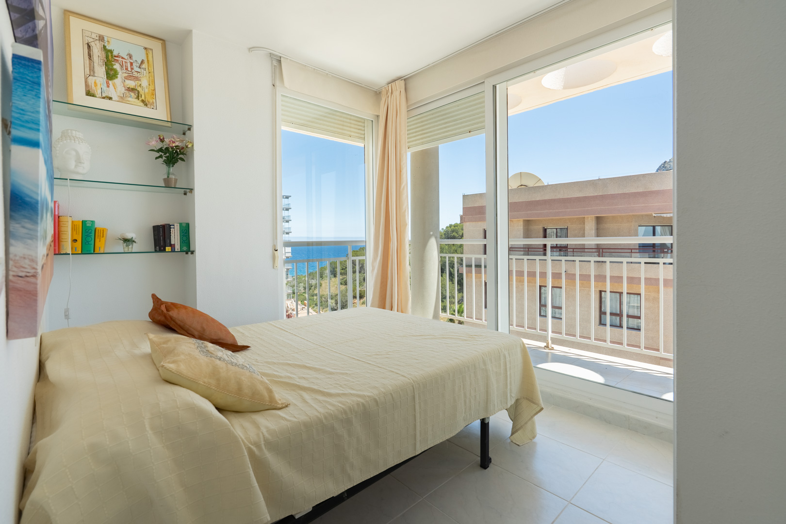 Chambre lumineuse avec lit double, étagères en verre intégrées et accès balcon offrant des vues partielles sur la mer et l'architecture.