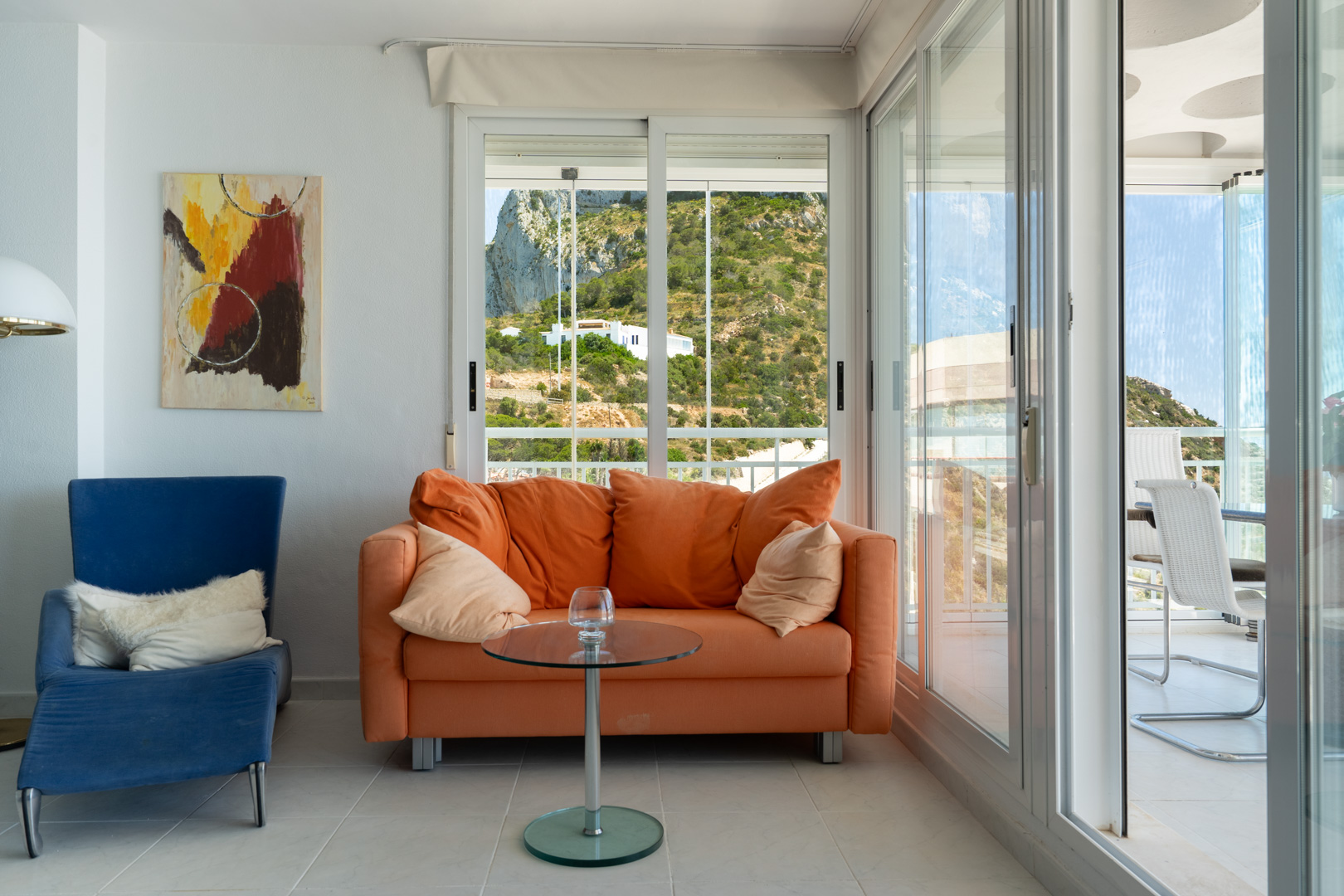 Coin salon confortable avec canapé orange, fauteuil bleu et œuvre d'art abstraite. De grandes fenêtres offrent une vue sur la colline verdoyante, menant à une terrasse couverte lumineuse.