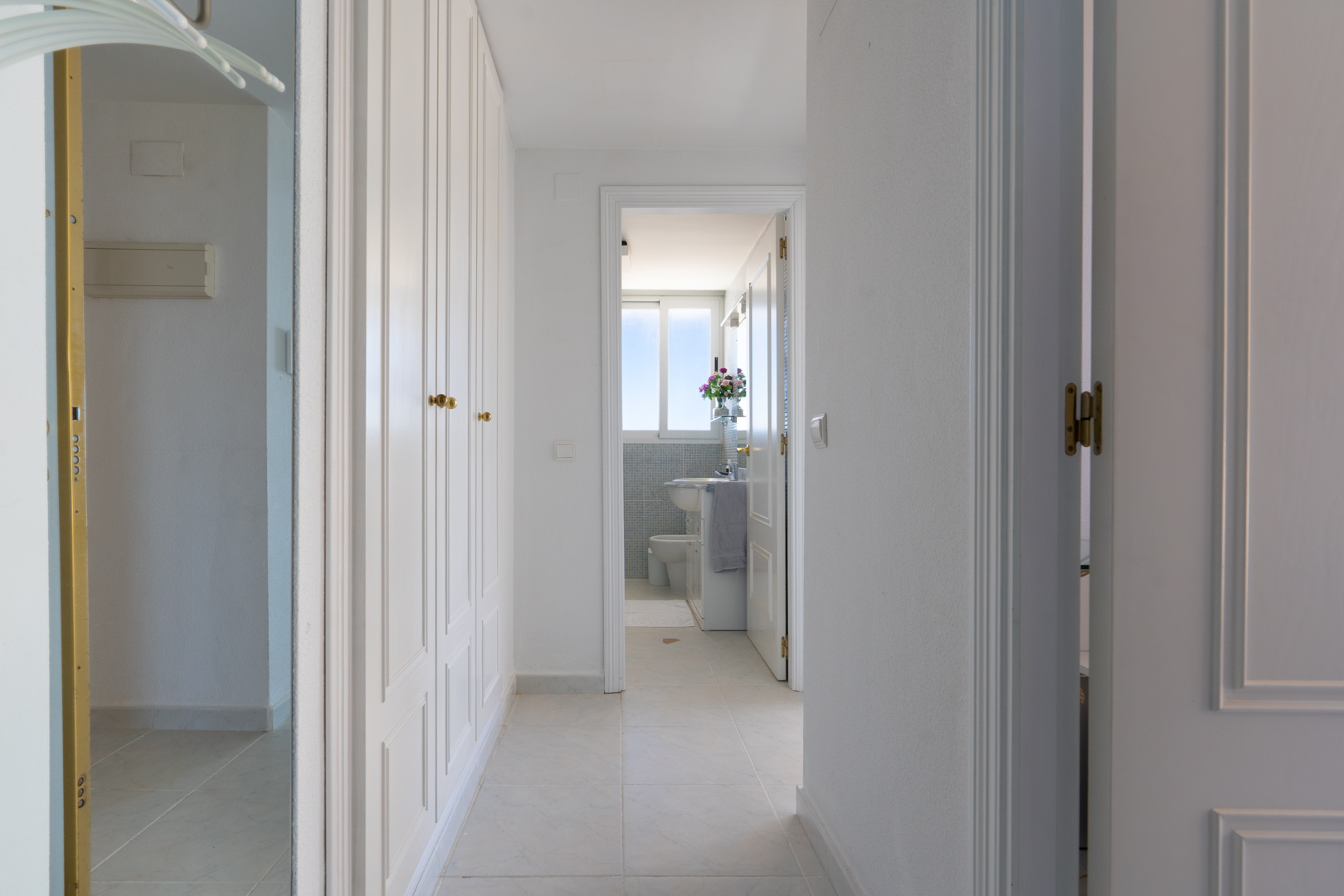 Couloir lumineux avec armoires encastrées blanches et poignées dorées. Le sol carrelé mène à une salle de bain visible par une porte ouverte, présentant une fenêtre et des carreaux de mosaïque bleus.