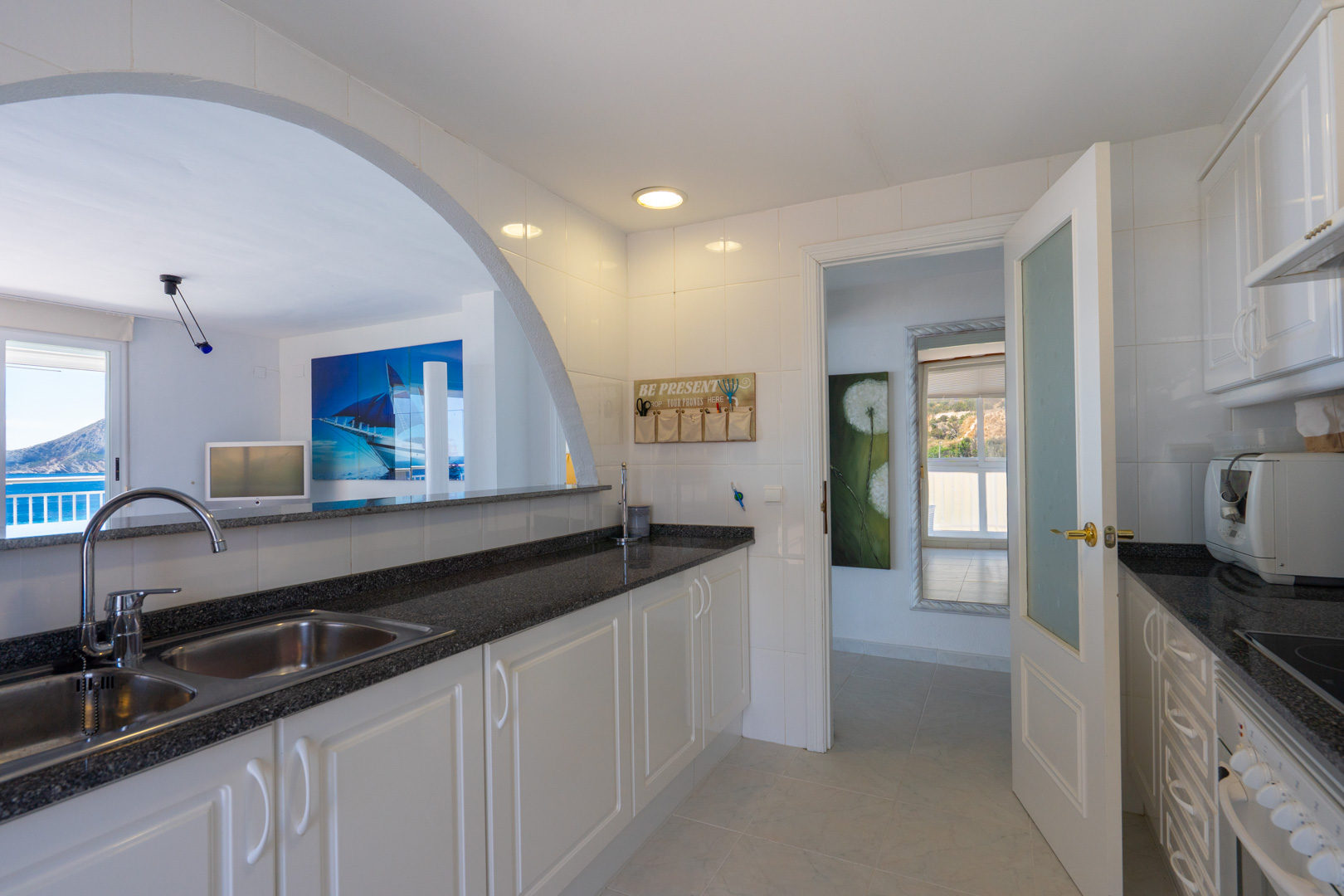 Cuisine lumineuse aux murs carrelés blancs, plan de travail foncé et double évier, reliée par une arche à un espace de vie avec vue sur mer et montagne.