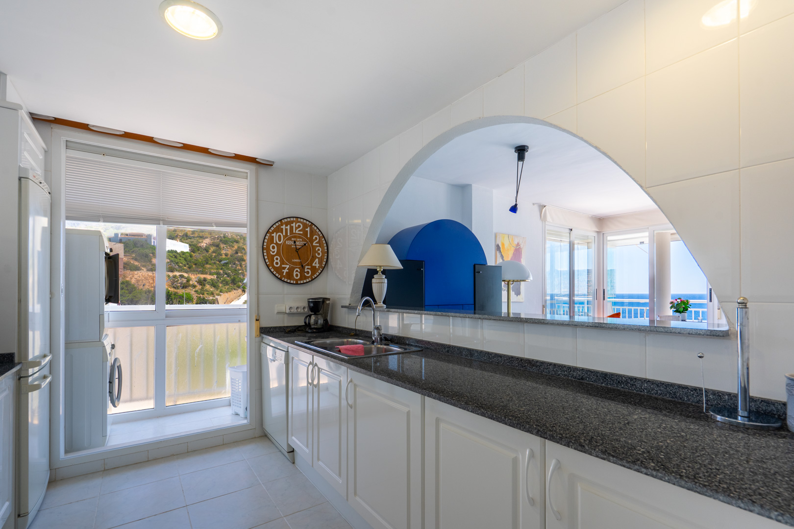 Cuisine semi-ouverte avec des armoires blanches, un plan de travail foncé et un espace buanderie intégré, reliée par une arche au salon offrant une vue sur la mer.