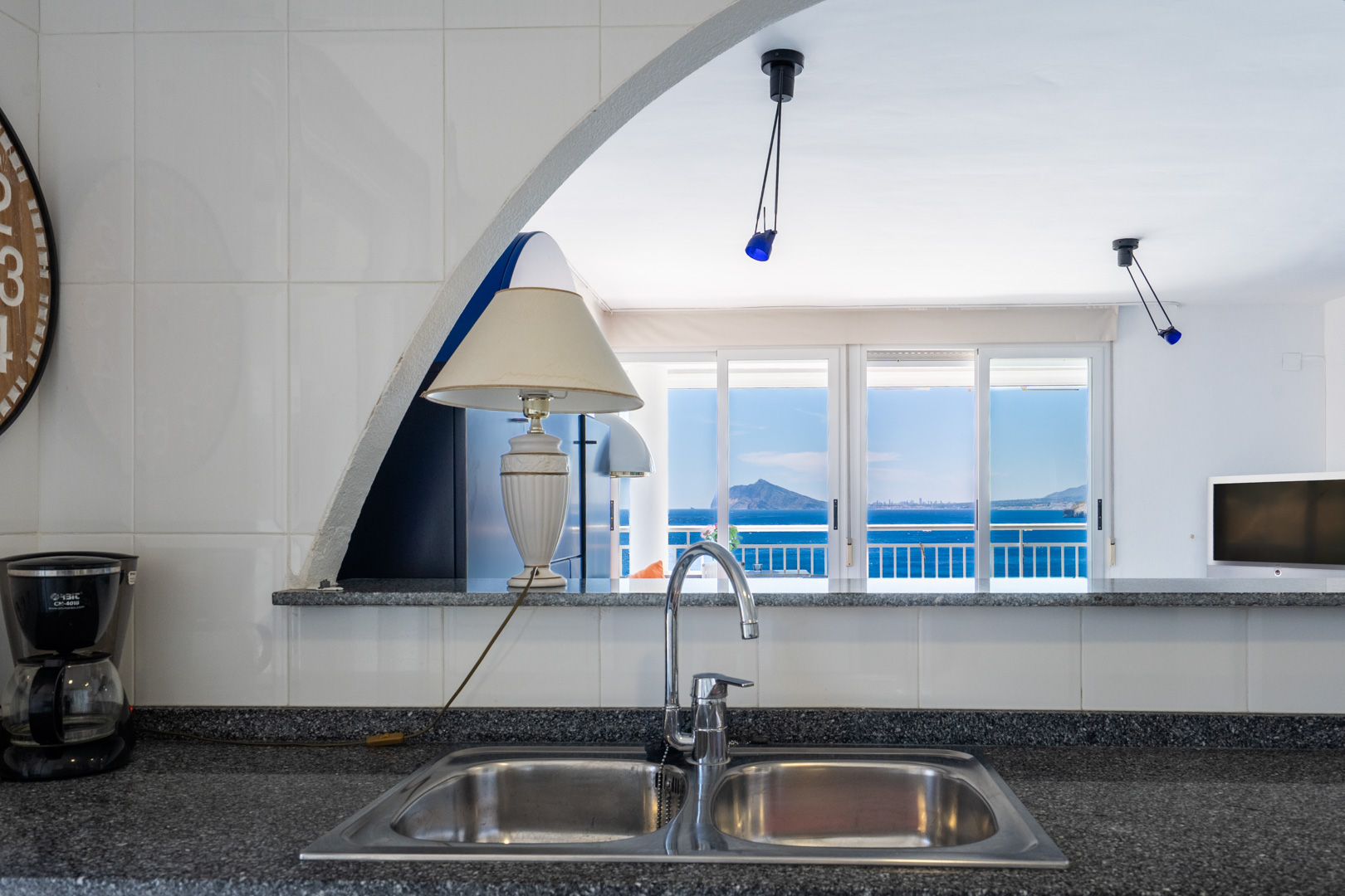 Espace cuisine avec évier double en acier inoxydable et plan de travail moucheté, encadrant des vues sur la mer et les montagnes lointaines à travers une arche et des portes coulissantes.