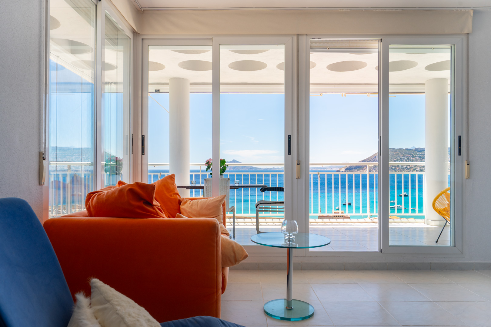 Espace de vie avec canapé orange vif et table d'appoint en verre, s'ouvrant sur une terrasse avec balustrade blanche et vue panoramique sur la mer turquoise.