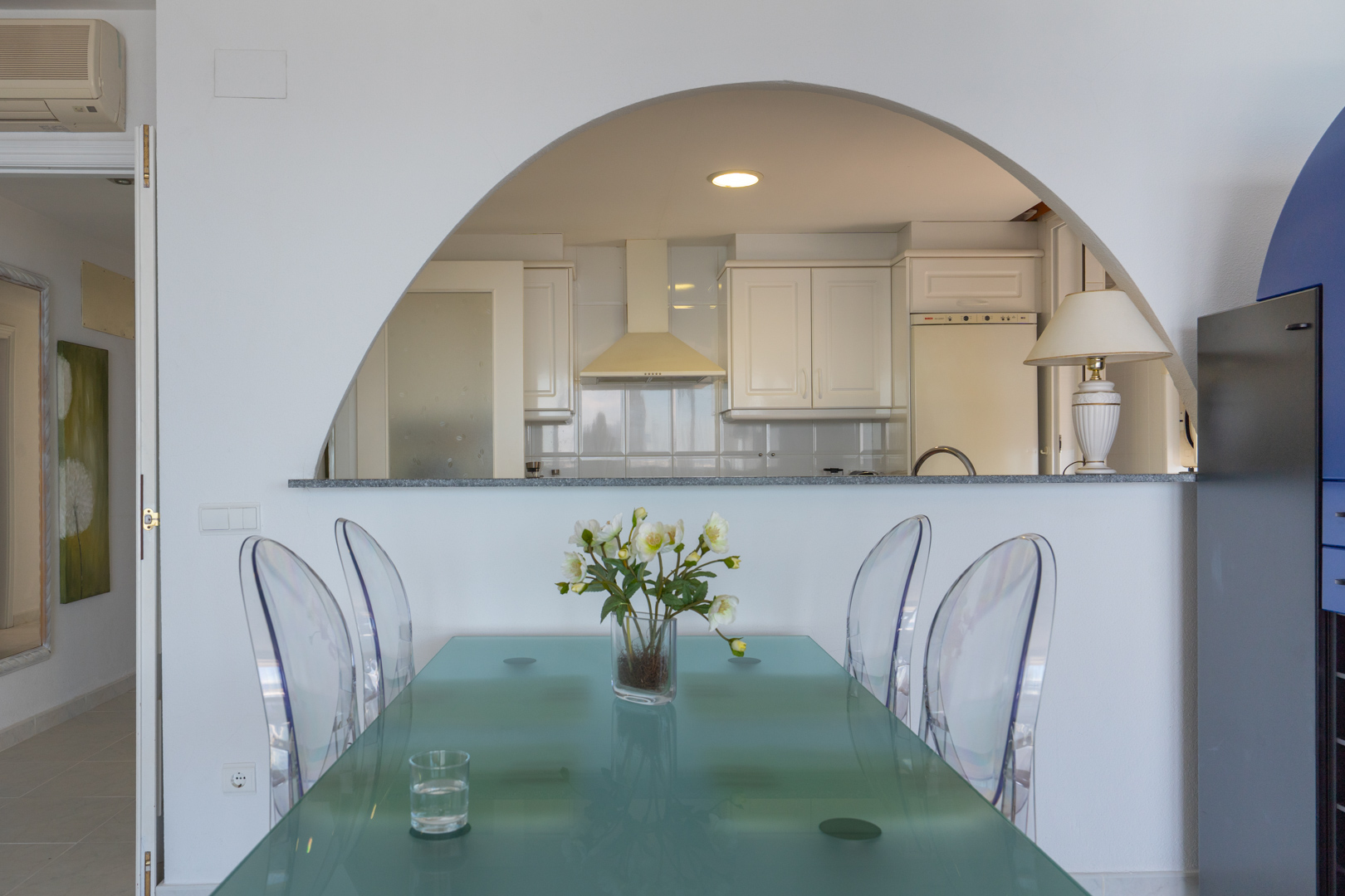 Espace repas avec table en verre moderne et chaises transparentes, ouvert sur une cuisine semi-ouverte par une arche, avec armoires blanches.
