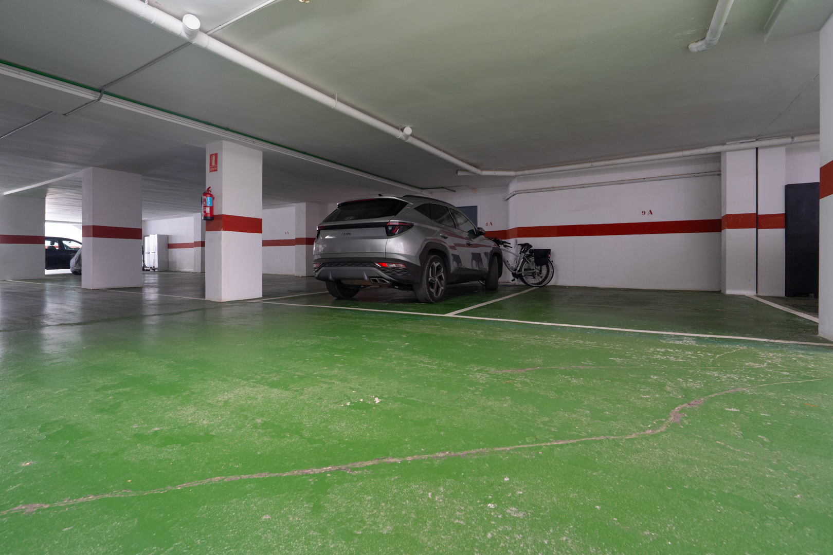 Place de parking privée dans un garage souterrain éclairé, avec sol en résine verte, piliers blancs à rayures rouges et extincteur. Un SUV gris est garé à côté d'un vélo.