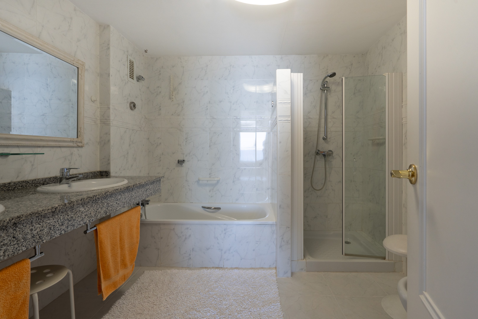 Salle de bain principale avec carrelage mural effet marbre, plan vasque en granit, baignoire et cabine de douche vitrée séparée.