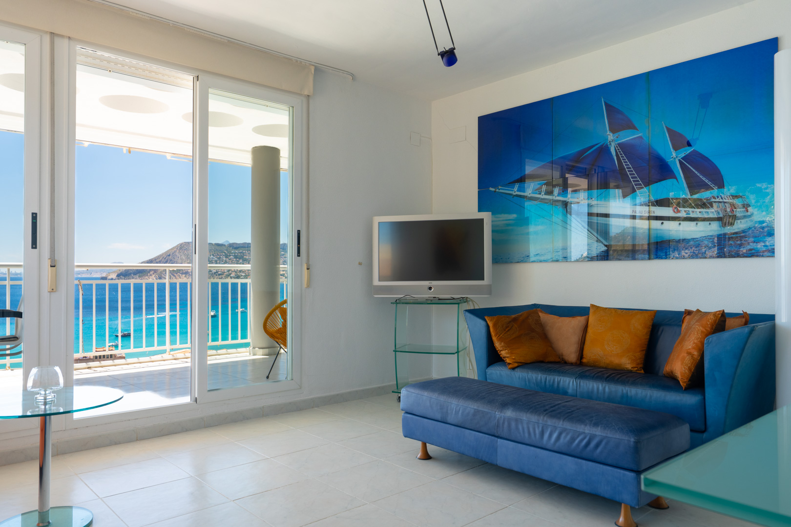 Salon lumineux avec canapé bleu, œuvre d'art marine et portes coulissantes s'ouvrant sur une terrasse offrant une vue panoramique sur la mer.