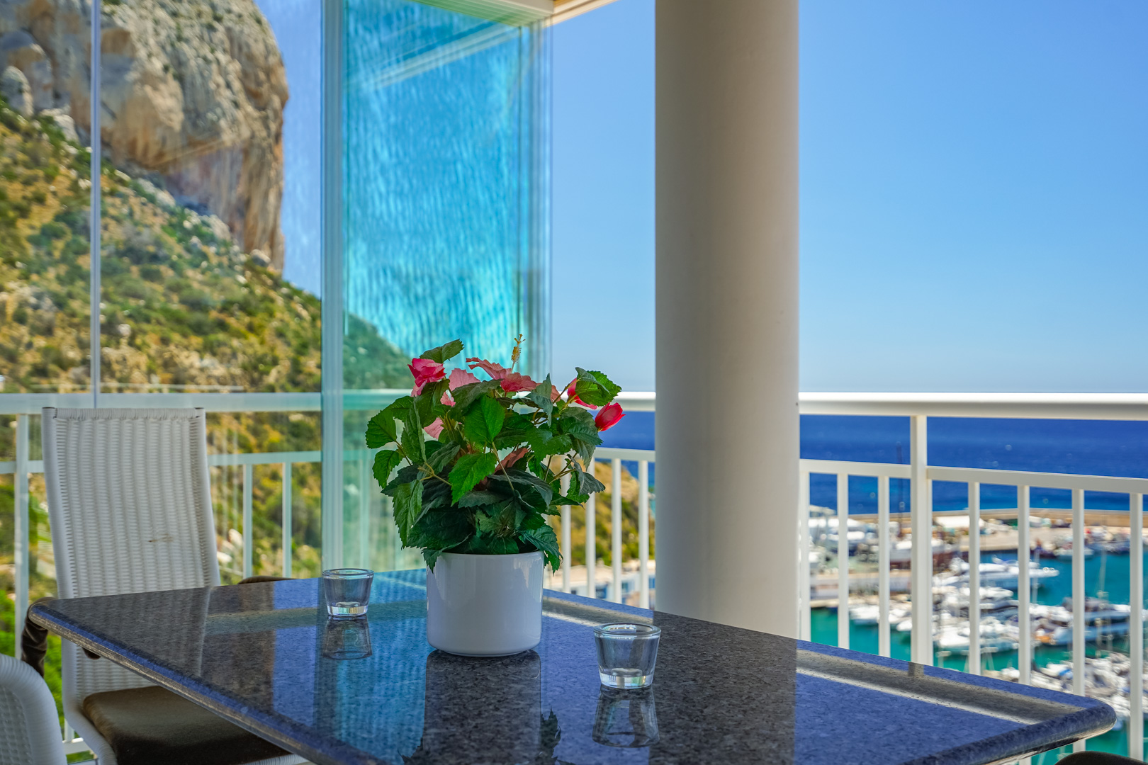 Terrasse avec table en granit, plante fleurie et panneaux vitrés, offrant des vues sur une montagne rocheuse et la marina.