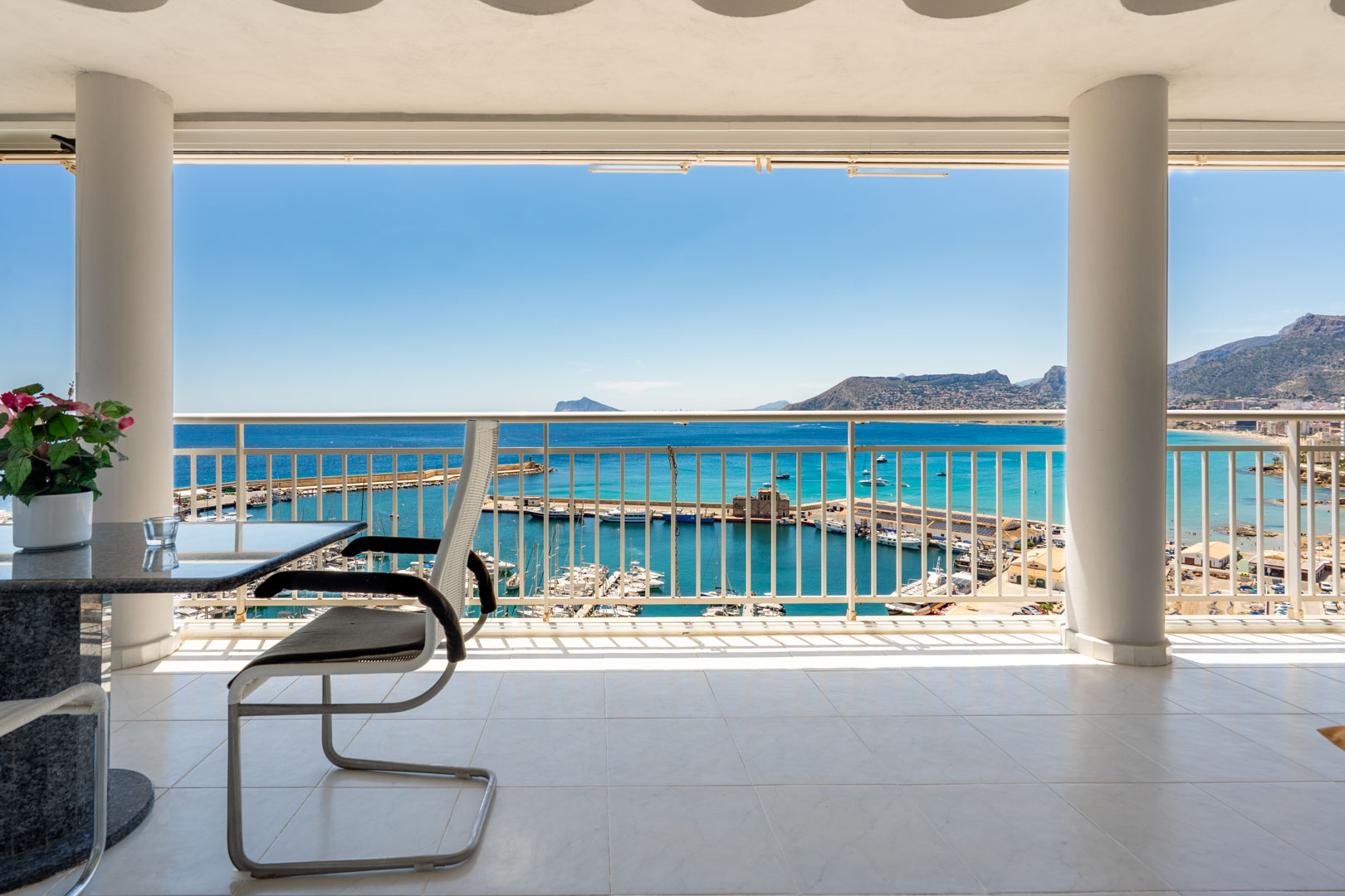Terrasse couverte avec carrelage blanc et colonnes structurelles, offrant une vue panoramique directe sur le port de plaisance de Calpe, la mer Méditerranée et les montagnes lointaines.