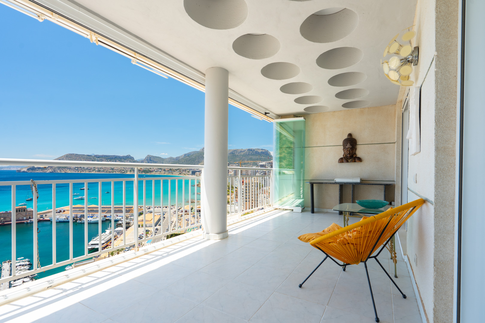 Vaste terrasse couverte avec carrelage blanc et plafond à motifs circulaires. Offre des vues panoramiques sur le port de plaisance, la mer turquoise et les montagnes lointaines, avec parois vitrées.