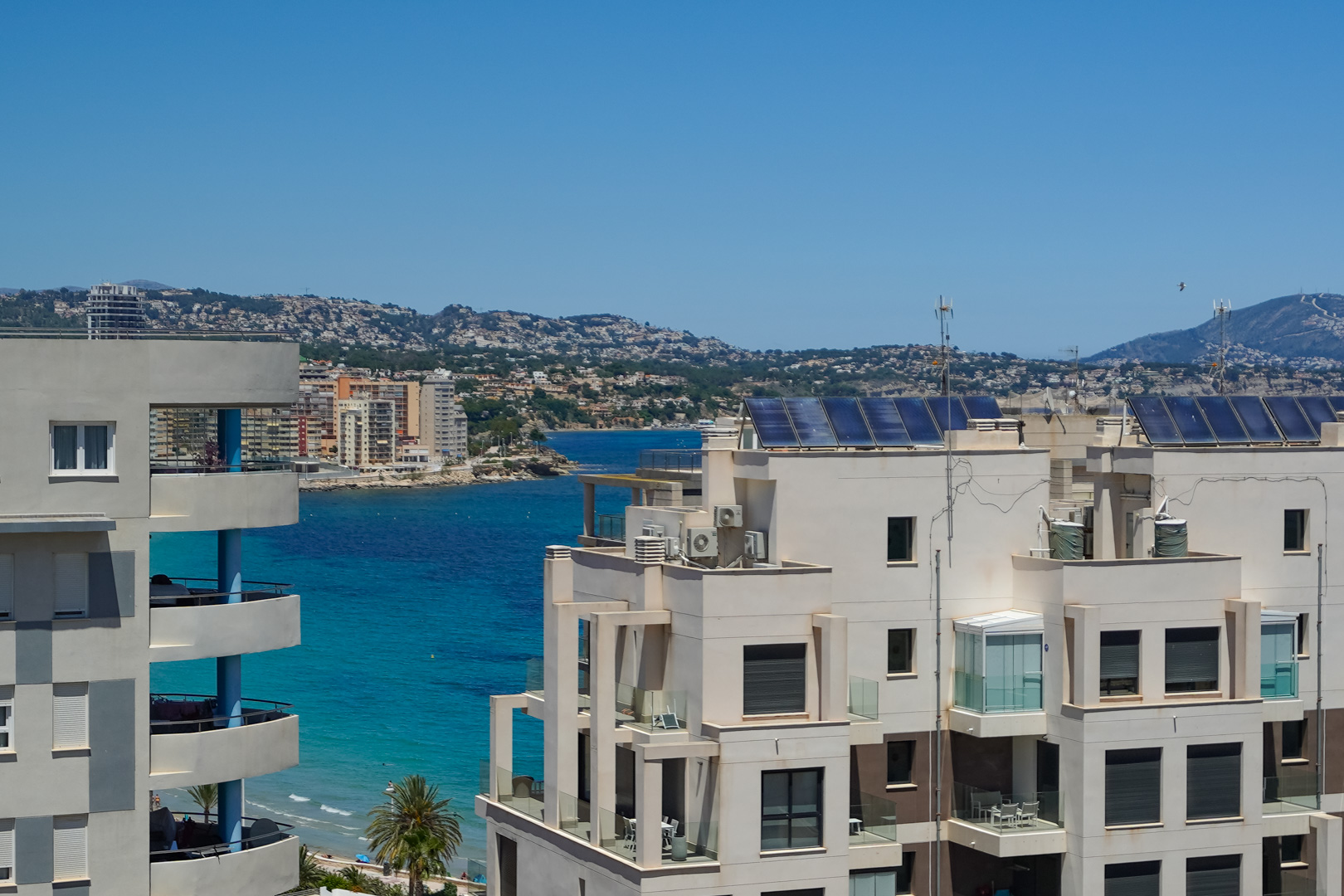 Vue élevée sur des bâtiments côtiers modernes avec balcons, surplombant la mer turquoise, la côte lointaine et les montagnes sous un ciel clair. Panneaux solaires visibles sur les toits.