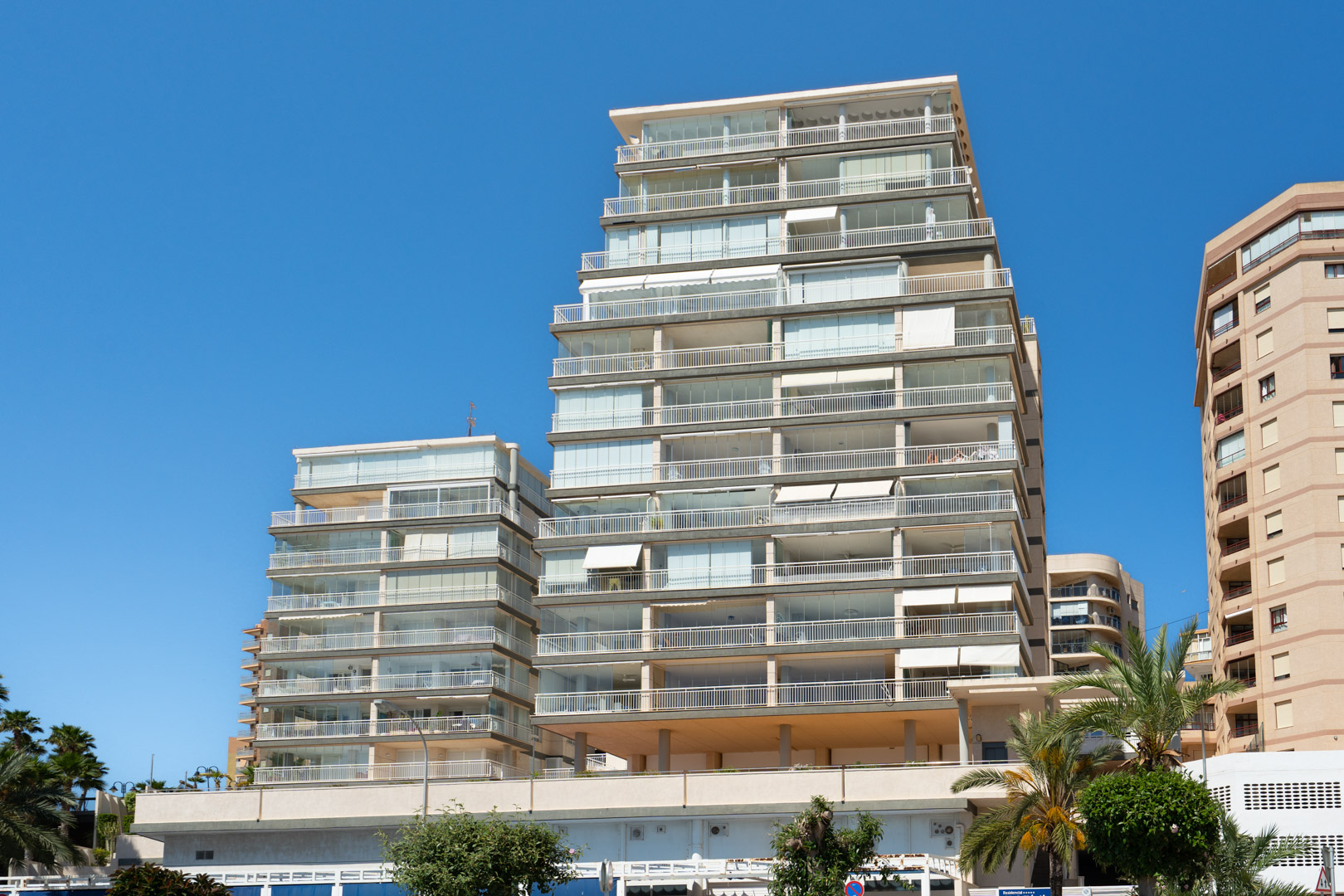 Vue extérieure d'un immeuble résidentiel moderne à plusieurs étages avec de nombreux balcons, dont plusieurs sont fermés par des rideaux de verre et des balustrades blanches, sous un ciel bleu. Des palmiers sont visibles.