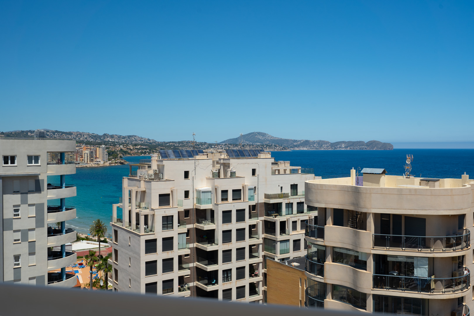 Vue mer élevée depuis une terrasse, présentant une architecture côtière moderne, la Méditerranée éclatante et des montagnes lointaines. Piscine et palmiers visibles.