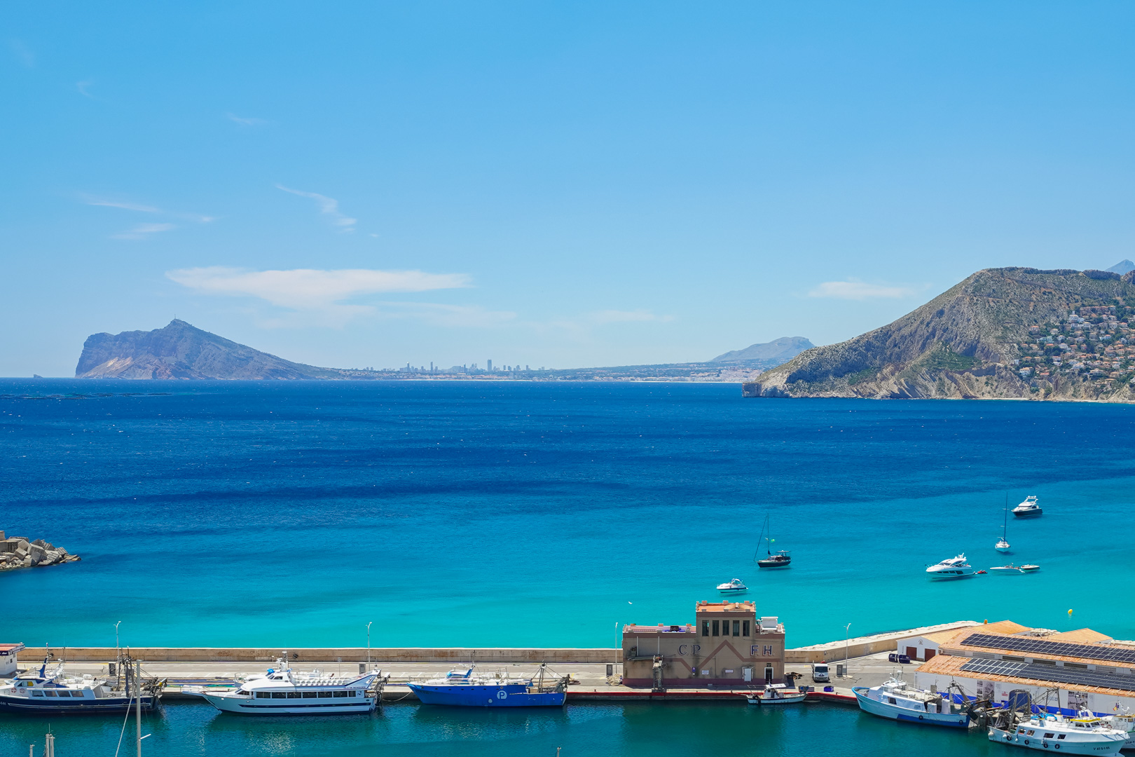 Vue panoramique sur le port de plaisance de Calpe, avec des bateaux, des eaux turquoise et des montagnes lointaines. Le Peñón de Ifach est visible à droite, avec un skyline à l'horizon.