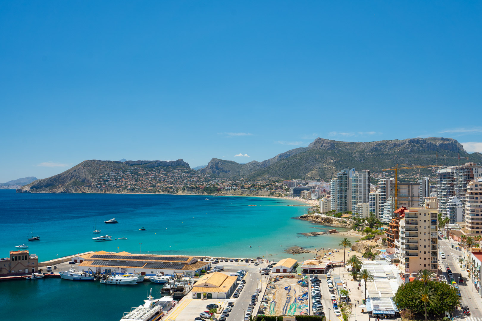 Vue panoramique sur le port de plaisance de Calpe, la baie turquoise et la Playa de La Fossa, encadrée par des montagnes lointaines sous un ciel clair.