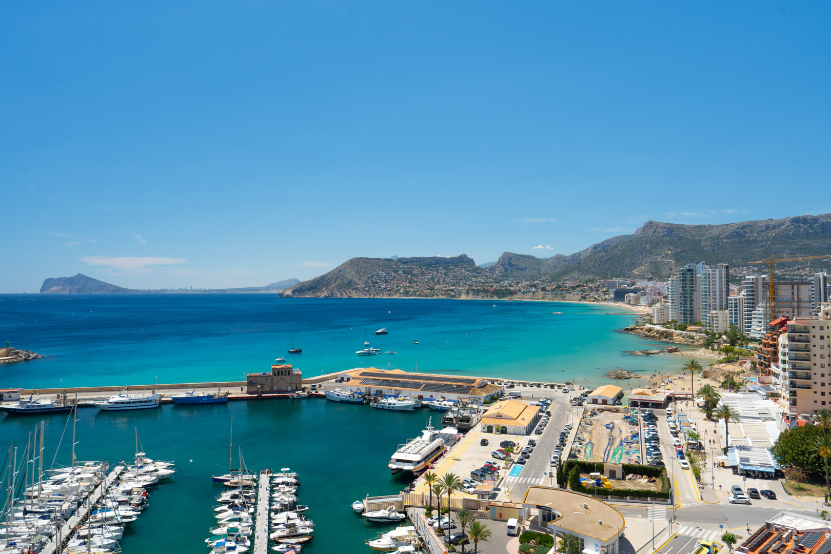 Vue panoramique sur le port de plaisance de Calpe, sa baie aux eaux turquoise, une plage de sable bordée de bâtiments côtiers et des montagnes lointaines.