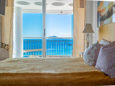 Chambre confortable avec lit et accès direct à un balcon, offrant des vues panoramiques sur la mer bleue, le port de plaisance et une île lointaine.