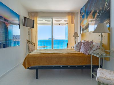 Chambre lumineuse avec tête de lit jaune et accès direct à une terrasse par des portes coulissantes en verre, offrant une vue étendue sur la mer bleue et une terre lointaine.