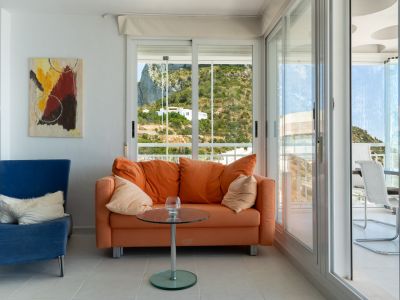 Coin salon confortable avec canapé orange, fauteuil bleu et œuvre d'art abstraite. De grandes fenêtres offrent une vue sur la colline verdoyante, menant à une terrasse couverte lumineuse.