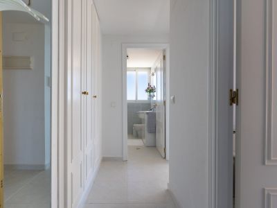 Couloir lumineux avec armoires encastrées blanches et poignées dorées. Le sol carrelé mène à une salle de bain visible par une porte ouverte, présentant une fenêtre et des carreaux de mosaïque bleus.