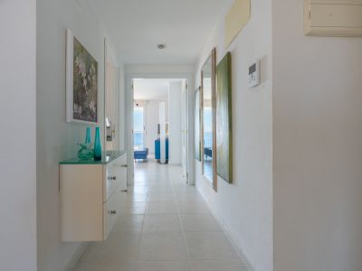 Couloir lumineux carrelé menant à un salon ensoleillé avec vue sur la mer. Comprend une console flottante en verre, des vases décoratifs et des œuvres d'art murales.