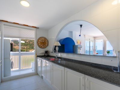 Cuisine semi-ouverte avec des armoires blanches, un plan de travail foncé et un espace buanderie intégré, reliée par une arche au salon offrant une vue sur la mer.