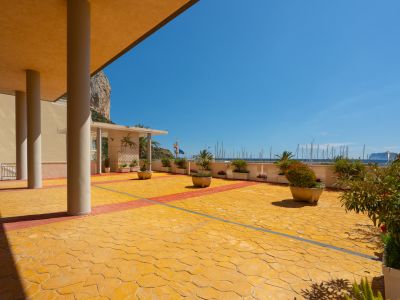 Espace extérieur commun avec pavage texturé jaune et rouge, plantes en pot, et vues directes sur la marina avec mâts de bateaux et l'emblématique Peñón de Ifach.