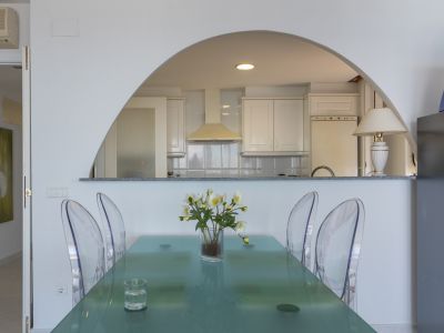 Espace repas avec table en verre moderne et chaises transparentes, ouvert sur une cuisine semi-ouverte par une arche, avec armoires blanches.