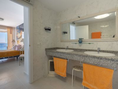 Salle de bain attenante avec double vasque en granit et carrelage effet marbre blanc. Par l'embrasure, on aperçoit la chambre adjacente avec vue sur la mer.
